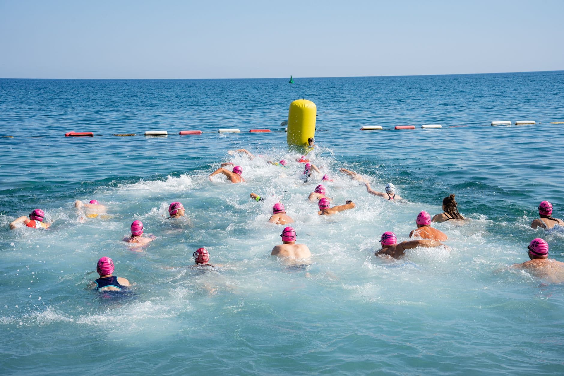 Field of open-water swimmers in pink caps heading out towards a yellow turn buoy